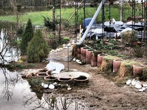 Garden by a flooded yard with circular brick planters filled with soil, a small evergreen shrub, and red brick edging; water covers the ground around the beds and a white tarp is visible in the background.